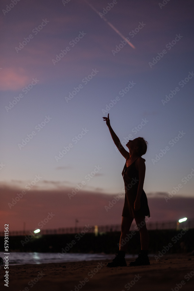 Fototapeta premium Girl Reaching for the Stars on a Beach at Sunset With a Colorful Sky in the Background.
