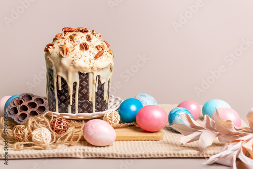 Traditional orthodox Easter cake - Paska with white chocolate and nuts on white round plate, blue and pink colored Easter eggs on light beige background in rustic style.
