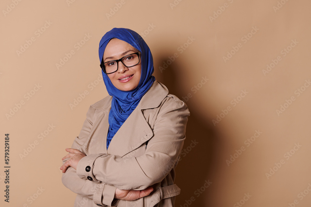 Portrait of a smiling middle-aged Muslim woman in elegant strict outfit and blue hijab, wearing spectacles, posing with arms crossed over isolated beige background. people. Middle-Eastern pretty women Portrait of a smiling middle-aged Muslim woman in elegant strict outfit and blue hijab, wearing spectacles, posing with arms crossed over isolated beige background. people. Middle-Eastern pretty women