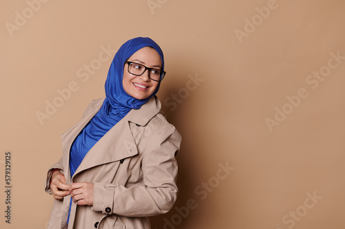 Stylish elegant confident Muslim woman in beige classic coat  blue hijab and black eyeglasses  smiling looking asideat a copy space on isolated beige background. Islamic female fashion and lifestyle