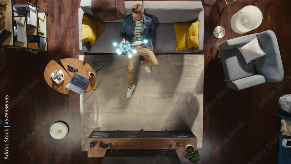 Top View Of Caucasian Man In the Loft Apartment Sitting Down on The Couch and Connecting Smartphone to Convenient Smart Home System. VFX Animation Visualizing Connected Devices. Laptop, TV, Speaker.