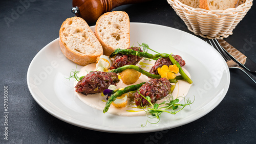 Cold appetizer of beef tartare with with asparagus, herbs, sauce and slices of bread.