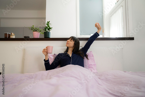 pretty young woman stretching in bed drinking coffee or tea with laptop teleworking at home stretching when waking up in the morning
