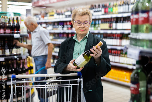 mature woman with glasses chooses bottle of wine in alcohol section of supermarket
