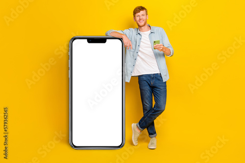 Photo of cheerful positive man wear jeans shirt typing modern gadget empty space isolated yellow color background