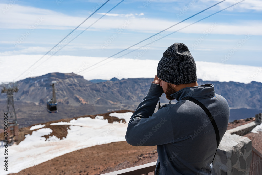Joven hombre turista con ropa de invierno fotografiando teleférico en paisaje rocoso con nieve, vistas desde el teide