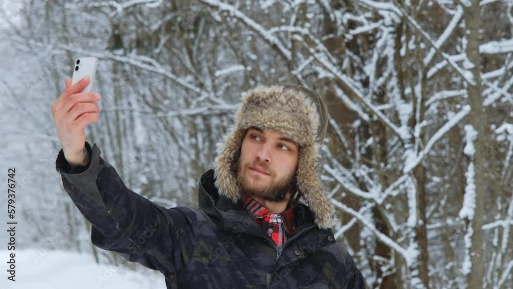 Young Caucasian male blogger in fluffy hat with earflaps stands in winter forest and takes selfie video or photo on phone. Friendly charismatic guy makes various grimaces on his face. Young Caucasian male blogger in fluffy hat with earflaps stands in winter forest and takes selfie video or photo on phone. Friendly charismatic guy makes various grimaces on his face.