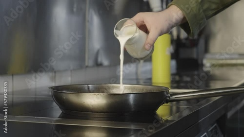 The chef pours milk and cream into a pan on the stove. Preparation of sauce in restaurant kitchen for dinner dishes.