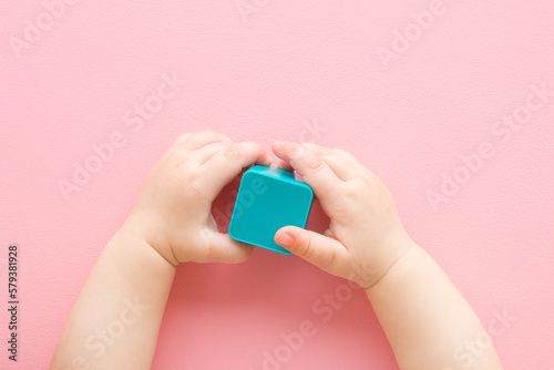 Baby girl hands holding and playing with light blue plastic shape on light pink table background. Pastel color. Closeup. Infant development toy. Point of view shot. Top down view.