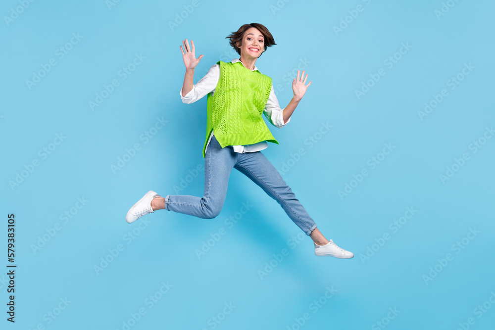 Full size photo of satisfied adorable girl with brown hairdo wear green vest jeans flying jumping isolated on blue color background