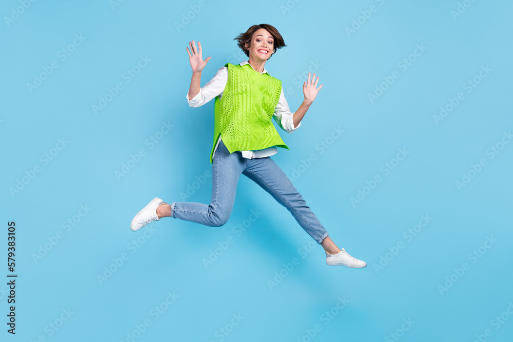 Full size photo of satisfied adorable girl with brown hairdo wear green vest jeans flying jumping isolated on blue color background Full size photo of satisfied adorable girl with brown hairdo wear green vest jeans flying jumping isolated on blue color background