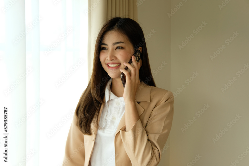 Happy young asian business woman wearing suit holding mobile phone standing in her workstation office Happy young asian business woman wearing suit holding mobile phone standing in her workstation office