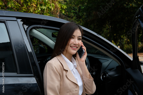 Happy confident professional woman posing near office building while using smartphone and opening car door. Young business female wearing a suit and a business bag. Successful businesswoman concept.