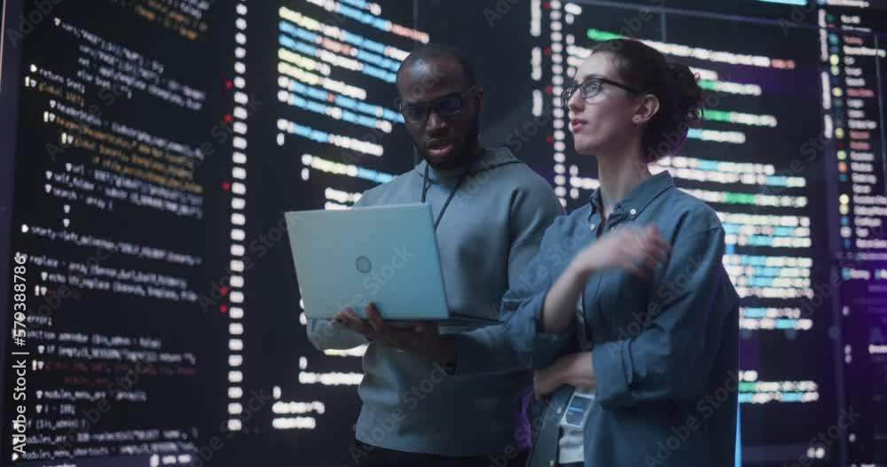 Portrait Two Diverse Developpers Using Laptop Computer, Discussing Lines of Code that Appear on Big Screens Surrounding Them. Male and Female Programmers Creating a Software Together, Fixing Bugs