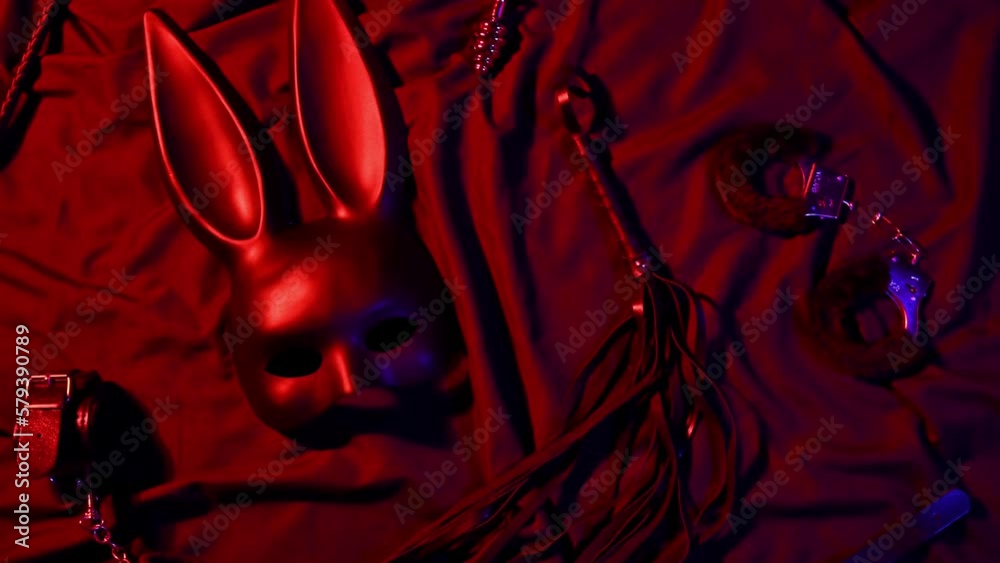 Erotic sex toys for adults for BDSM sex with submission and domination. Handcuffs, mask and leather flogger whip on a black background with red neon light