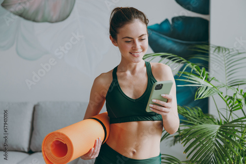 Fotografiet Young sporty athletic fitness trainer instructor woman wearing green tracksuit hold in hand yoga mat use mobile cell phone training do exercises at home gym indoor