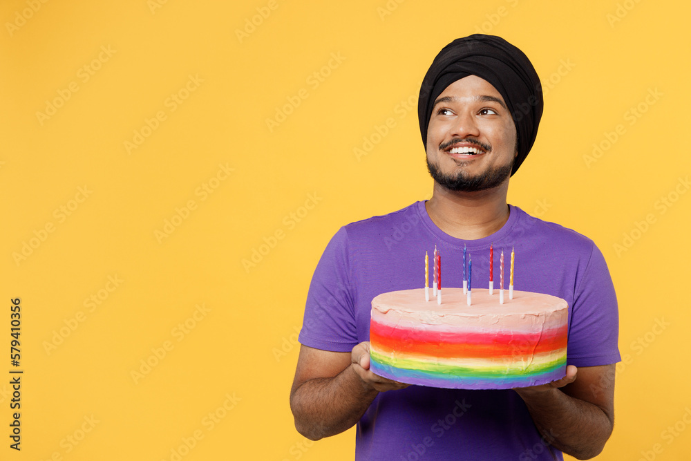 Smiling devotee Sikh Indian man ties his traditional turban dastar wears purple t-shirt hold in hand birthday cake with candles look aside on area isolated on plain yellow background studio portrait. Smiling devotee Sikh Indian man ties his traditional turban dastar wears purple t-shirt hold in hand birthday cake with candles look aside on area isolated on plain yellow background studio portrait.