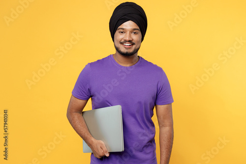 Fun devotee Sikh Indian IT man ties his traditional turban dastar wear purple t-shirt hold use work on laptop pc computer isolated on plain yellow background studio portrait. People lifestyle concept. photo
