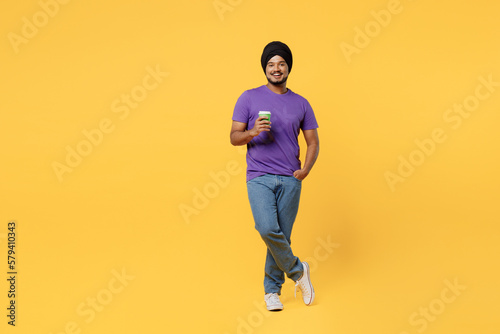 Full body devotee Sikh Indian man ties his traditional turban dastar wear purple t-shirt hold takeaway delivery craft paper brown cup coffee to go isolated on plain yellow background studio portrait.
