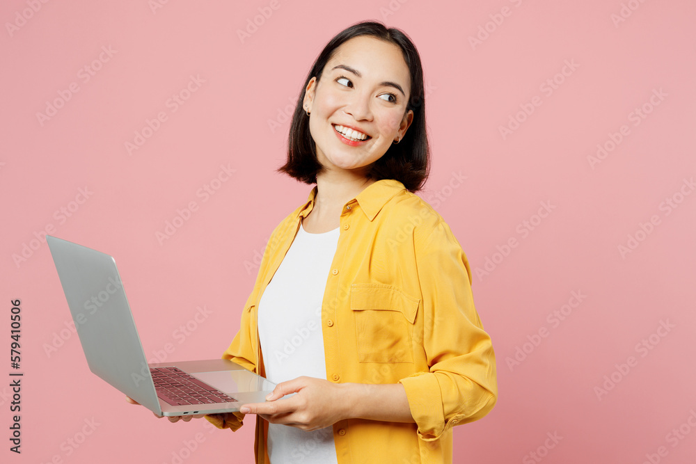 Young IT woman of Asian ethnicity wears yellow shirt white t-shirt hold use work on laptop pc computer look aside on area isolated on plain pastel pink background studio portrait. Lifestyle concept. Young IT woman of Asian ethnicity wears yellow shirt white t-shirt hold use work on laptop pc computer look aside on area isolated on plain pastel pink background studio portrait. Lifestyle concept.