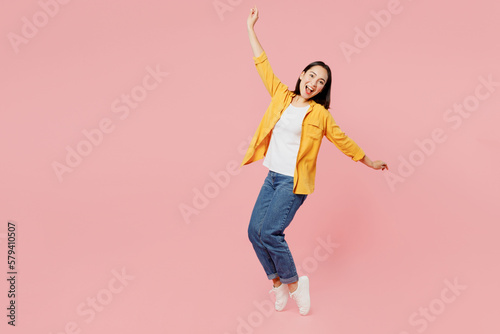 Full body side view young woman of Asian ethnicity wears yellow shirt white t-shirt leaning back with outstretched hands stand on toes isolated on plain pastel light pink background studio portrait.