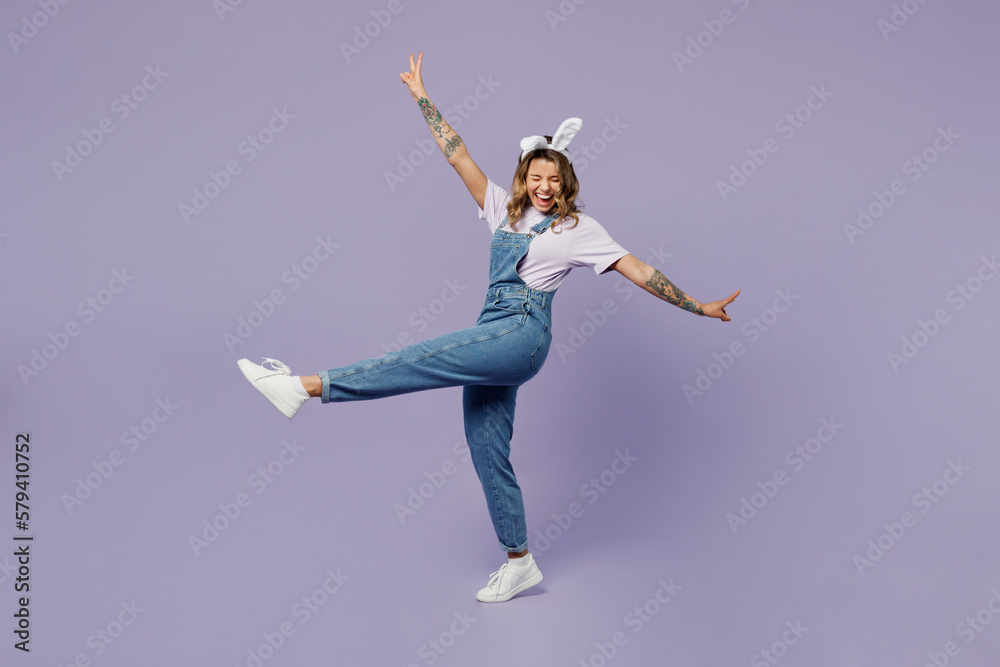 Full body side view young woman wear white casual clothes bunny rabbit ears stand with outstretched hands raise up leg isolated on plain pastel light purple background. Lifestyle Happy Easter concept.