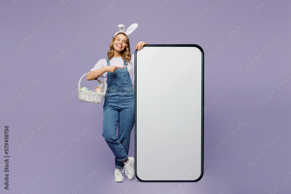 Full body young woman wear casual clothes bunny rabbit ears point index finger indicate on big huge blank screen mobile cell phone with area isolated on plain purple background. Happy Easter concept. - obrazy, fototapety, plakaty 