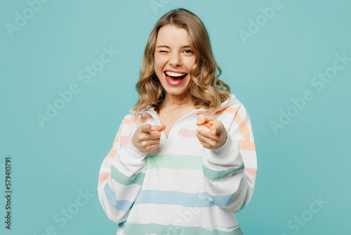 Young cheerful happy blonde woman wear hoody point index finger camera on you motivating encourage wink blink eye isolated on plain pastel light blue cyan background studio portrait Lifestyle concept