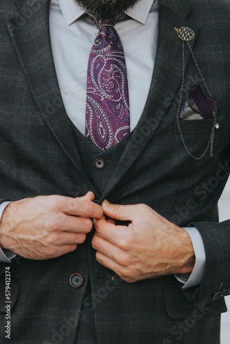 A man buttoning his gray check jacket partnered with purple paisley tie and a lapel chain