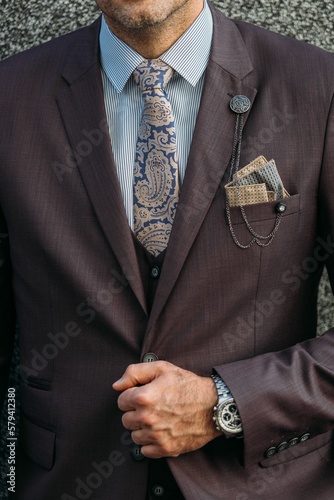 Man in a brown suit, white striped shirt with paisley tie and a lapel chain jewelry