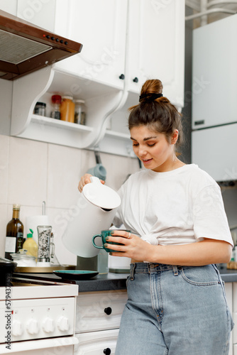 A brunette girl with hair styled in a bun is making tea in the kitchen in the morning. A girl in a white T-shirt and jeans is making tea in a white kitchen.