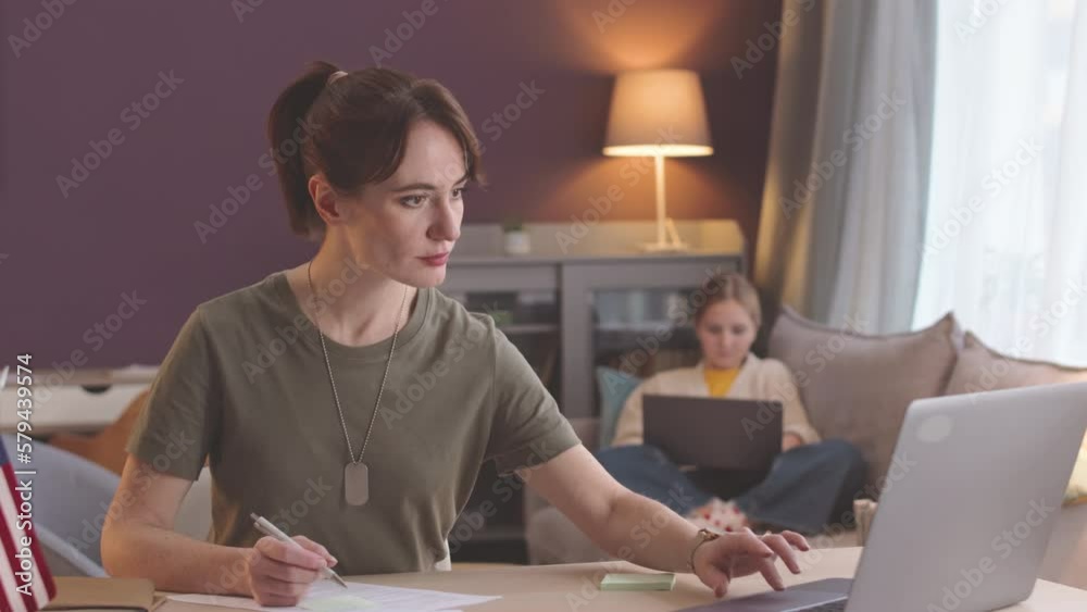 Waist up shot of Caucasian female army officer working on laptop from home while her preteen daughter surfing the Net on couch in background