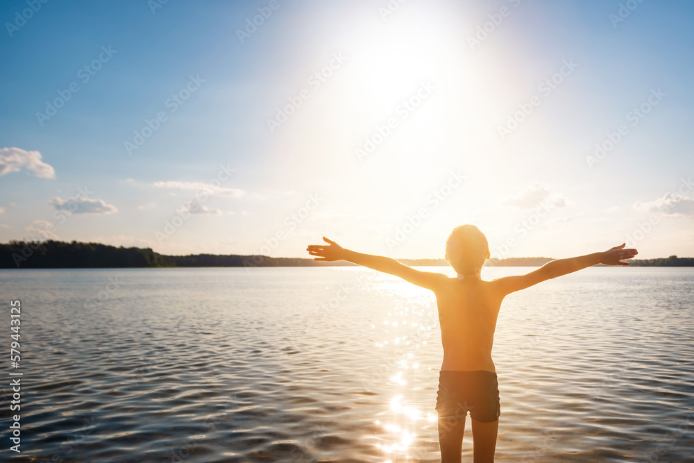 Fototapeta premium Shirtless child in warm sunset sunlight stands with his hands raised to the sun. Sun treatment, heliotherapy concept. Production of vitamin D in human cells.