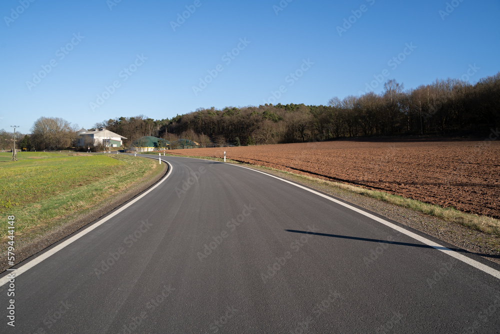 Asphalt road between grass and arable land with blue sunny sky in spring