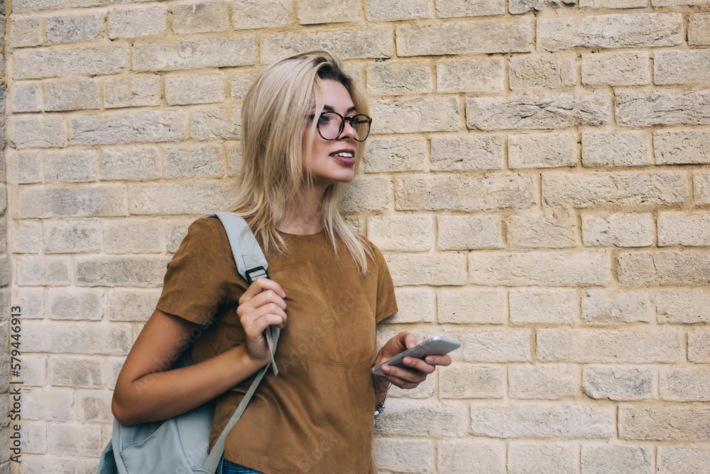 Fototapeta premium Smiling woman with handbag and smartphone standing near brick wall