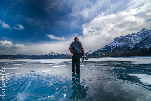 young man with beard in sweater walks on blue ice of abraham lake and holds in hands ice skates in jasper and banff national park