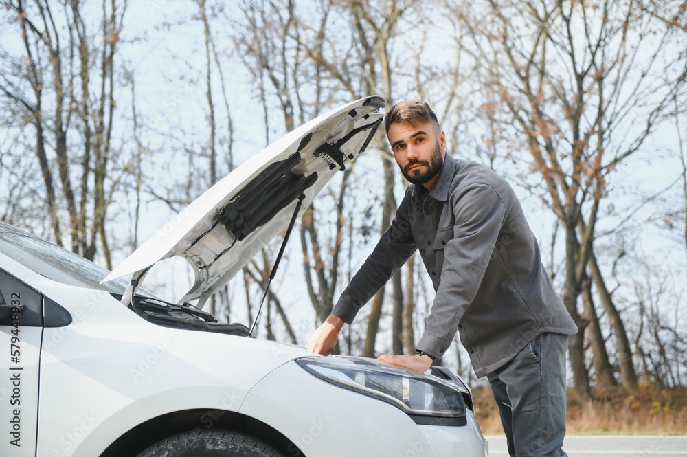 A young man near a broken car with an open hood on the roadside. A young man near a broken car with an open hood on the roadside.
