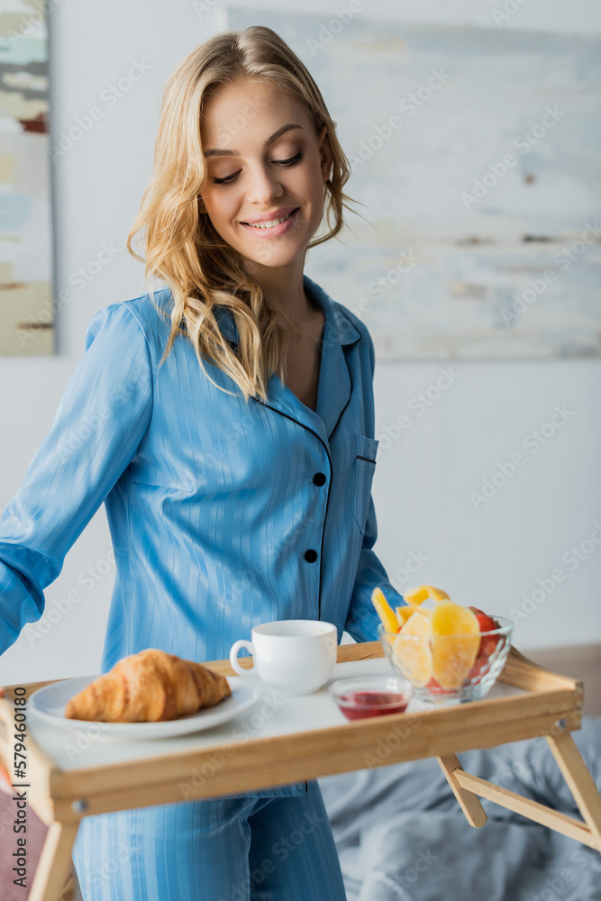smiling young woman in blue nightwear holding tray with breakfast in bedroom. smiling young woman in blue nightwear holding tray with breakfast in bedroom.