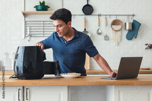 Handsome man standing at kitchen table and cooking toast bread healthy with recipe by Air Fryer machine, using laptop computer learning online cooking class in modern kitchen at home.