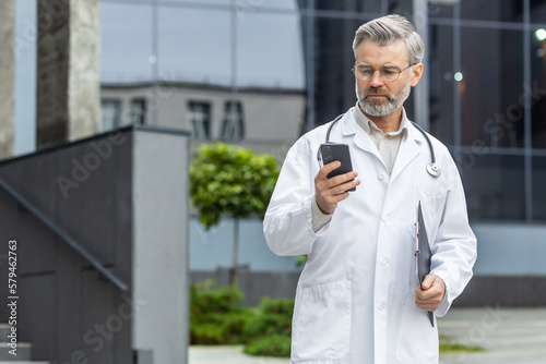 Foto An older gray-haired male doctor is standing outside the clinic in a uniform and holding a folder