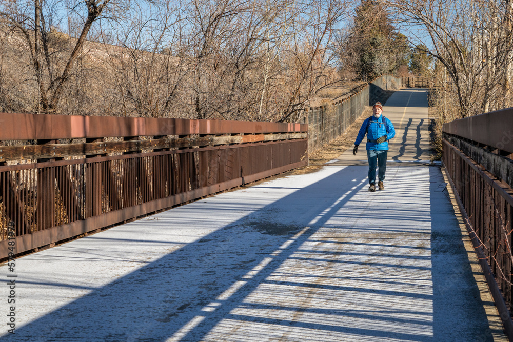 older man with a backpack is walking on a biking trail and footbridge covered by frost, winter morning in Fort Collins, Colorado