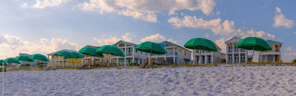 Bright green beach umbrellas on the white sandy shore in Destin Florida, Scenic panoramic landscape with multi-storey seafront homes against cloudy blue sky.