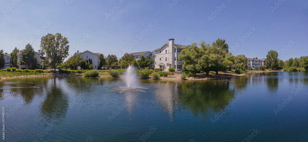 Beautiful apartments with view of a small lake with fountain in Boise Idaho. Scenic landscape of rental properties overlooking a reflective body of water against blue sky.