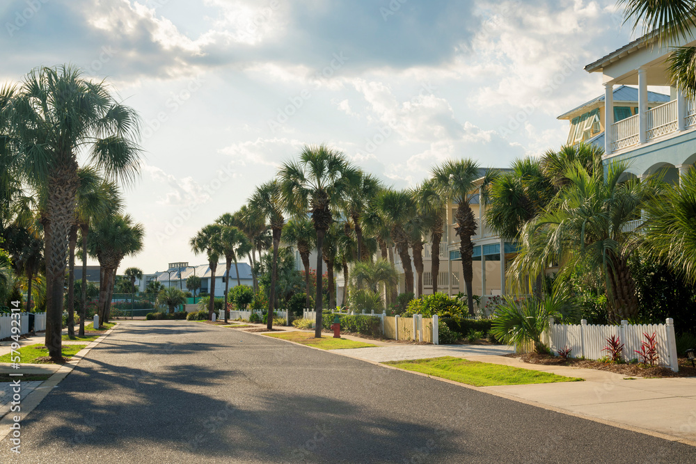 Street in a residential area with fenced houses and sidewalks along the trees in Destin, Florida. There is an asphalt road in the middle against the sky.
