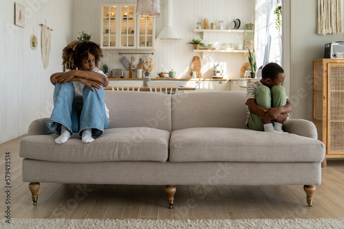 Offended African American mother and child boy sit on opposite sides of sofa pout at each other ignore silent after quarrel at home. Worried woman looks with displeasure at upset son, who turned away photo