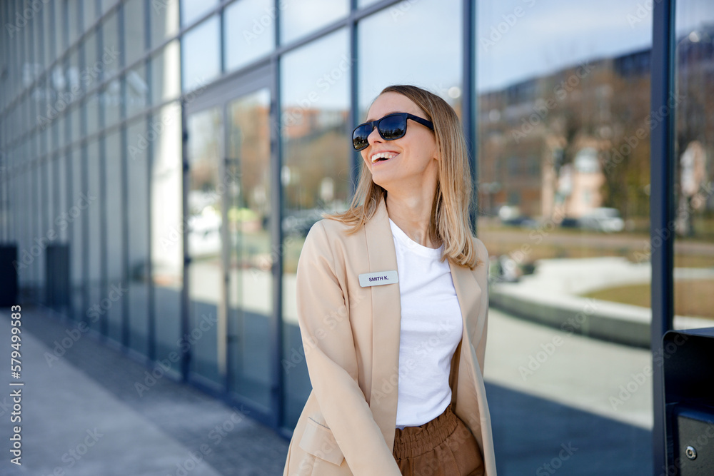 Happy manager smiling and looking at camera while walking near office center with crossed arms. Beautiful business woman wearing stylish formal jacket and trendy sunglasses.