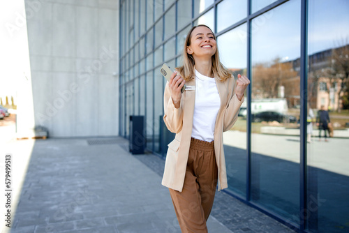 Excited businesswoman reading good news on smartphone while standing outdoors. Happy lady worker in suit making yes gesture with office center on background.