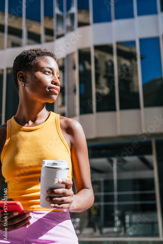 Young and attractive african woman with very short hair waiting in the street. She holds a reusable coffee mug and an smart phone.