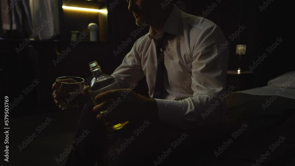 A man in a shirt sits on a bed at home in hotel, holds a bottle of alcohol, pours whiskey, scotch, cognac into a glass. Businessman drinks, sighs, tired after work, business meeting, relaxes A man in a shirt sits on a bed at home in hotel, holds a bottle of alcohol, pours whiskey, scotch, cognac into a glass. Businessman drinks, sighs, tired after work, business meeting, relaxes