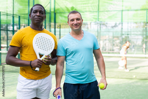 Portrait of positve men, african-american and caucasian, padel tennis players standing on outdoor court. © JackF